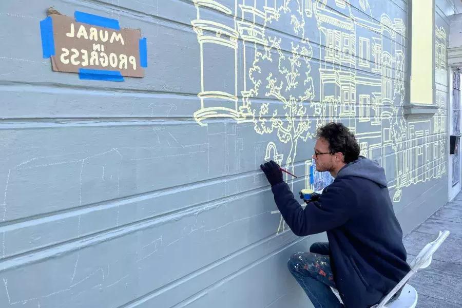An artist paints a mural on the side of a building in the mission district, with a sign taped onto the building that reads "Mural in Progress.San Francisco, CA.